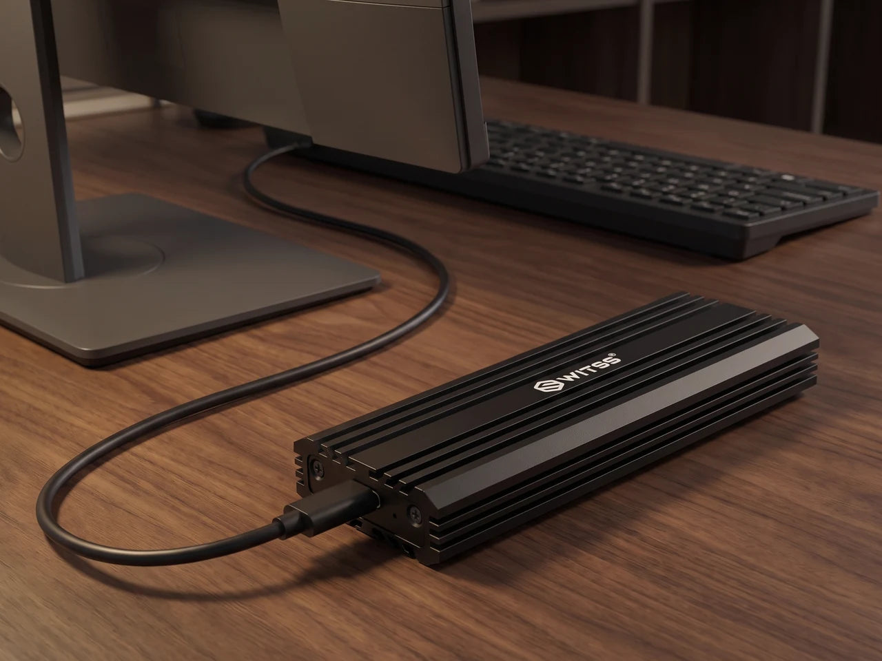 De Switss M.2 behuizing met de USB-C-kabel op een houten bureau, naast een beeldscherm en een toetsenbord