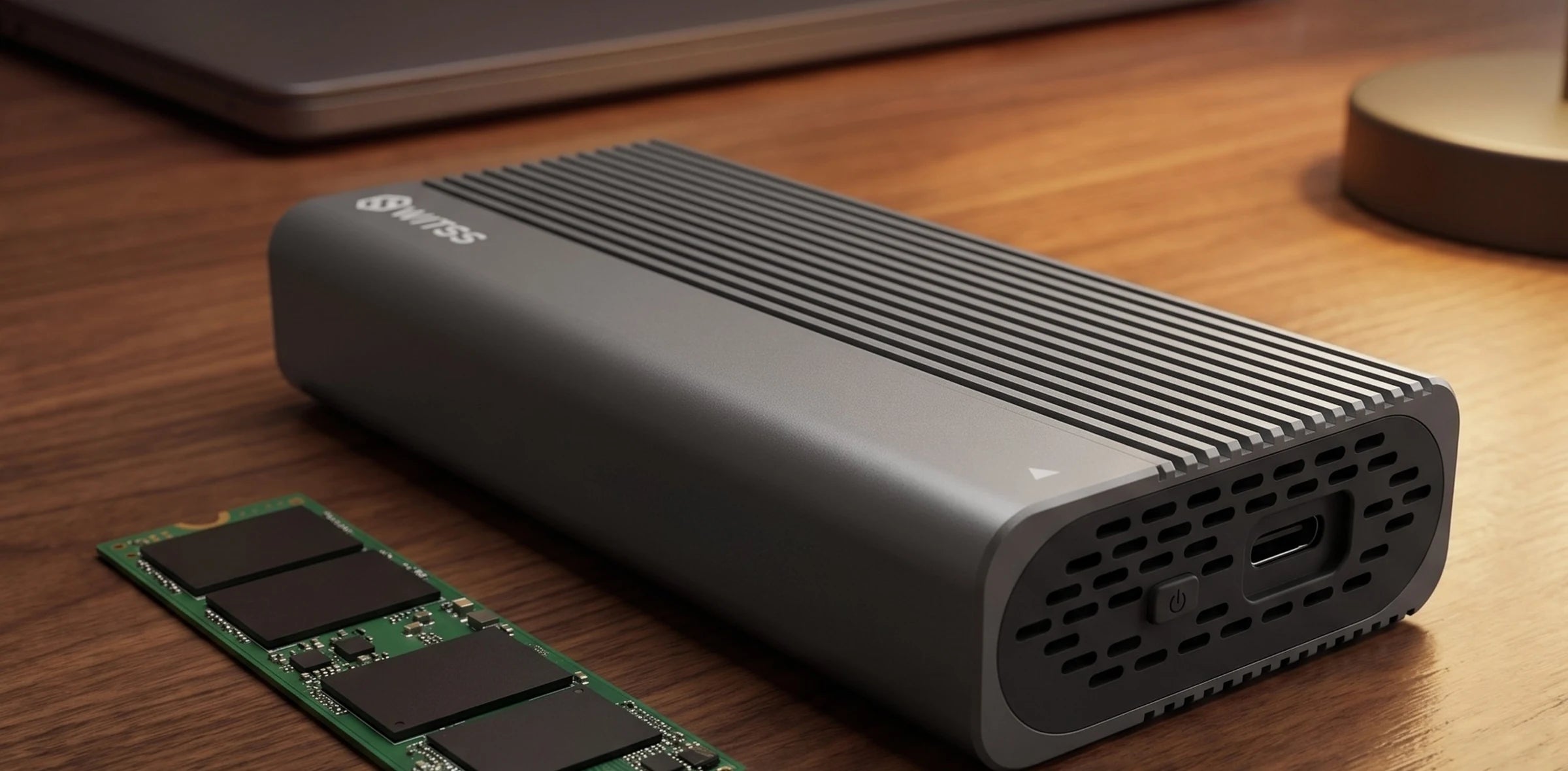 The Switss M.2 enclosure on a wooden desk, with a bare M.2 NVMe SSD beside it and a closed laptop in the background